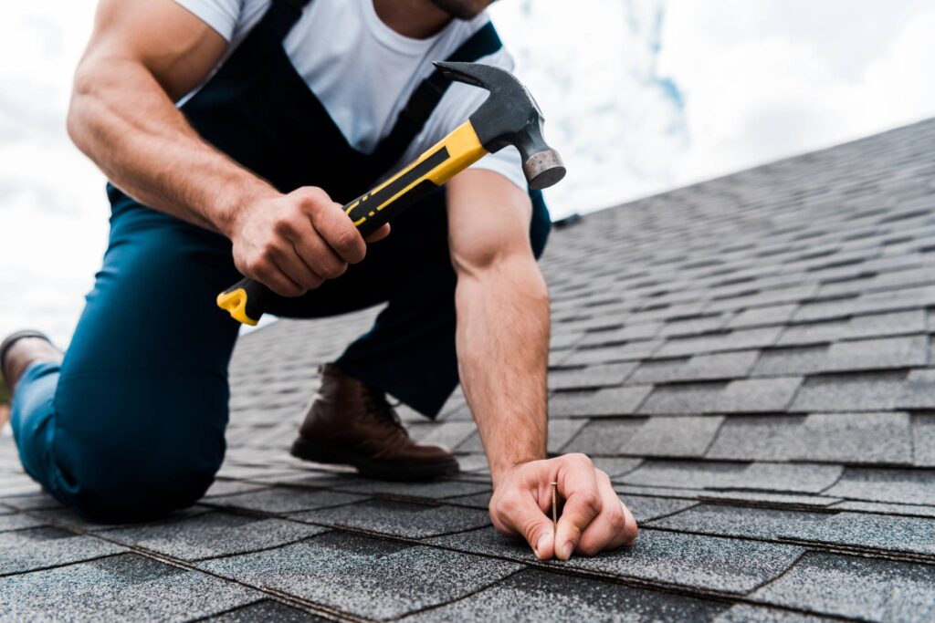 A man in a work uniform on a roof holding a hammer and nail performing roof repairs